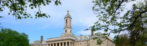 Stone university building with a tall clock tower framed by green tree branches against a blue sky.